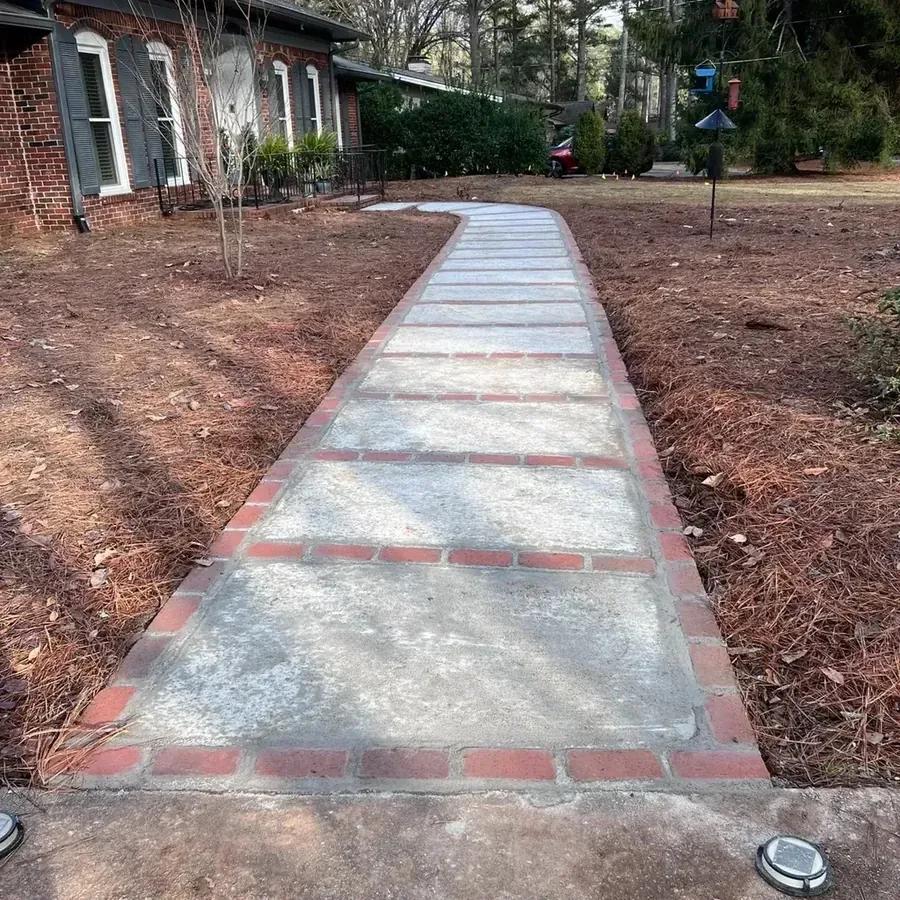 A brick ranch house with a concrete and brick front walkway between two bare pine-straw beds, under pine trees