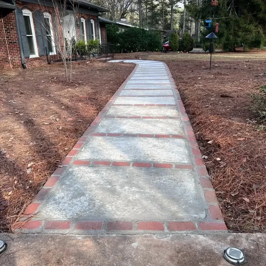 A brick ranch house with a concrete and brick front walkway between two bare pine-straw beds, under pine trees